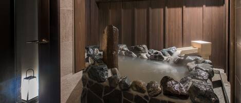 A stone hot tub with a wooden backdrop and a stone wall surrounding it.