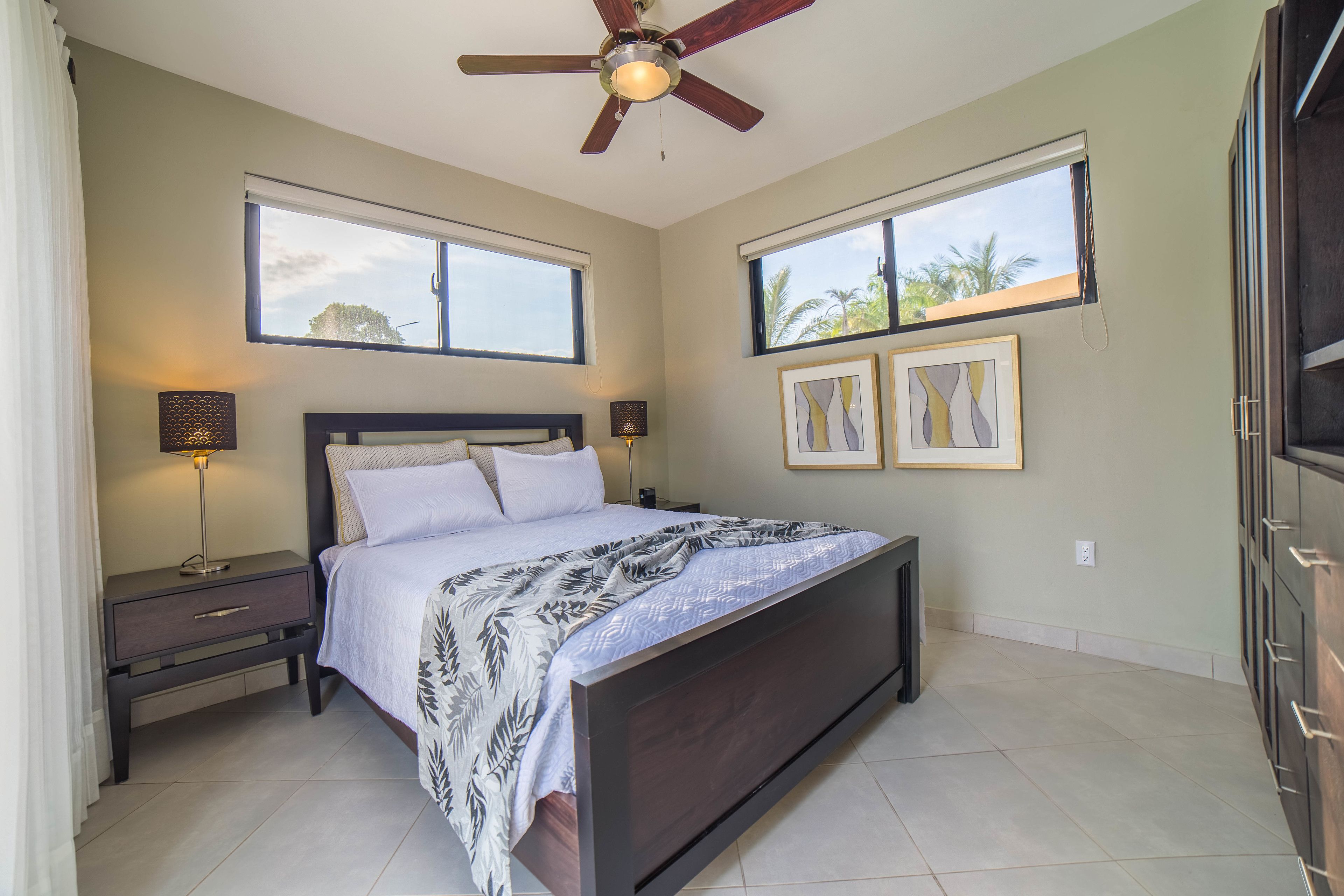 A bedroom with a bed, nightstands, a ceiling fan, two windows, and framed artwork on the wall.