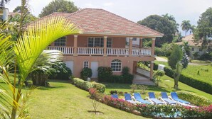 Une maison à deux étages avec un toit recouvert de tuiles rouges, un balcon et une pelouse bien entretenue agrémentée de chaises longues bleues.