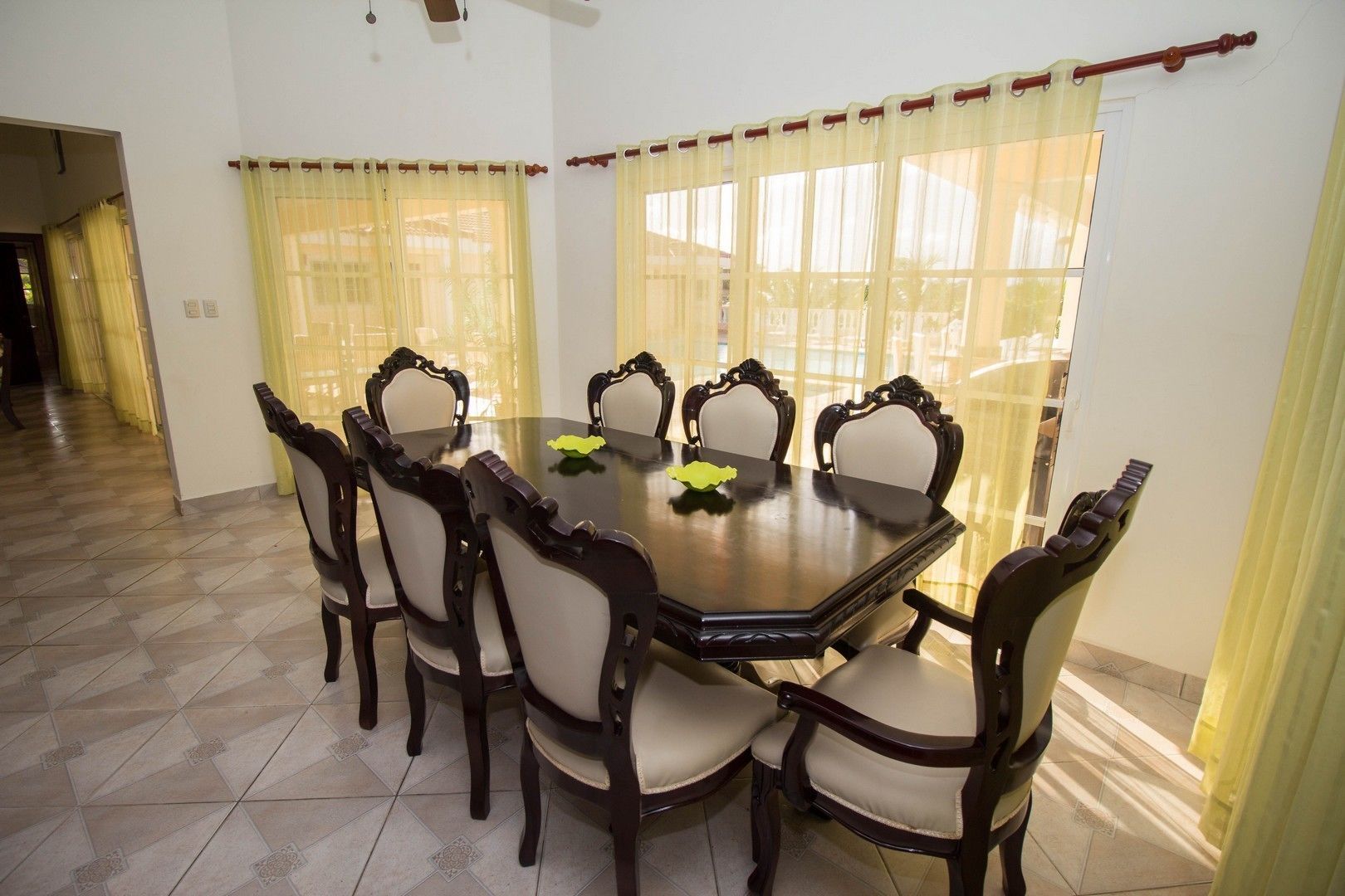 A dining area with a round table and six chairs, large windows with yellow curtains, and a tiled floor.