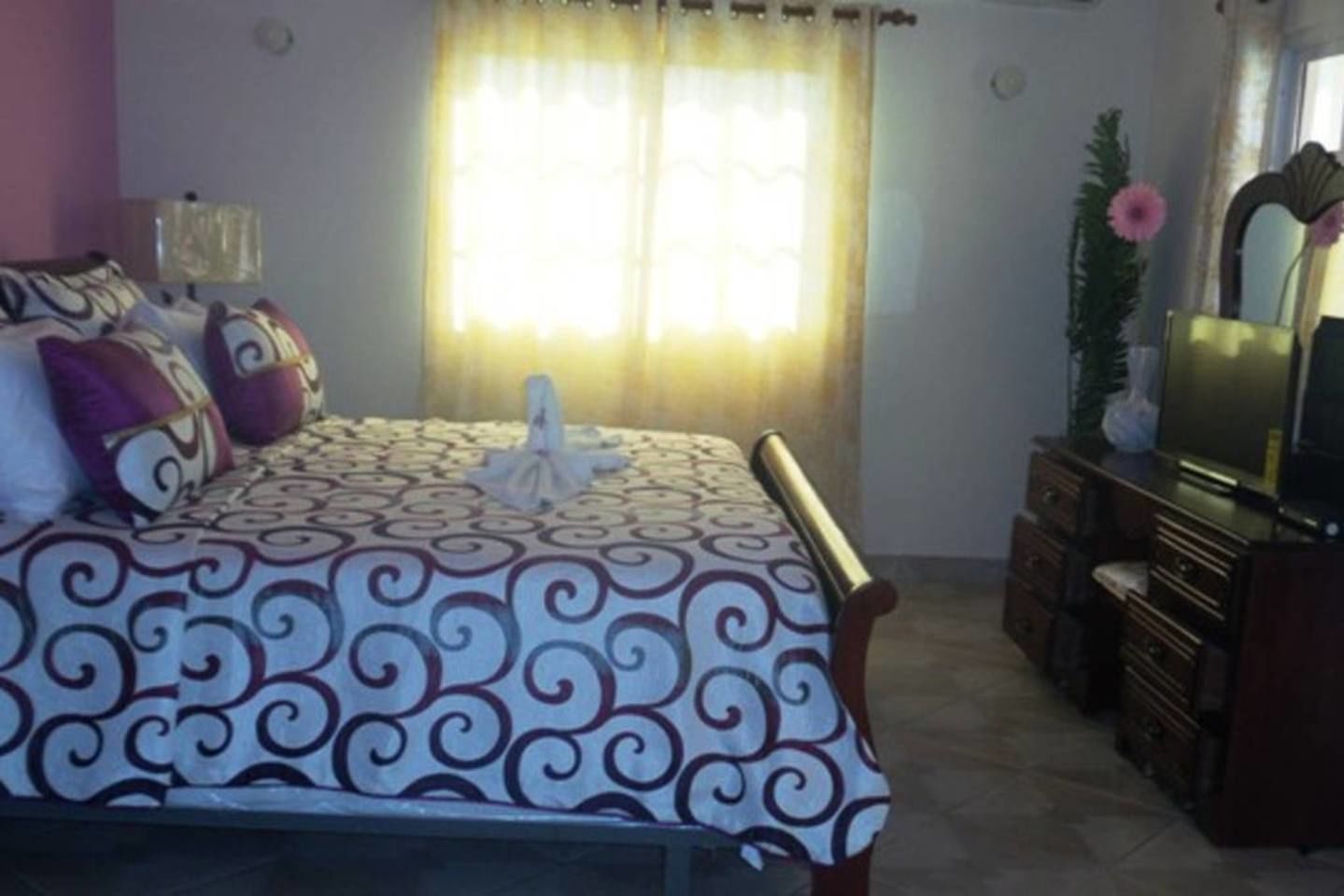 Une chambre à coucher avec un lit, une commode, un téléviseur et une fenêtre avec des rideaux.