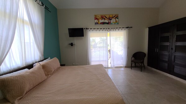 A bedroom with a bed, a chair, a TV, and a sliding glass door leading to a balcony.