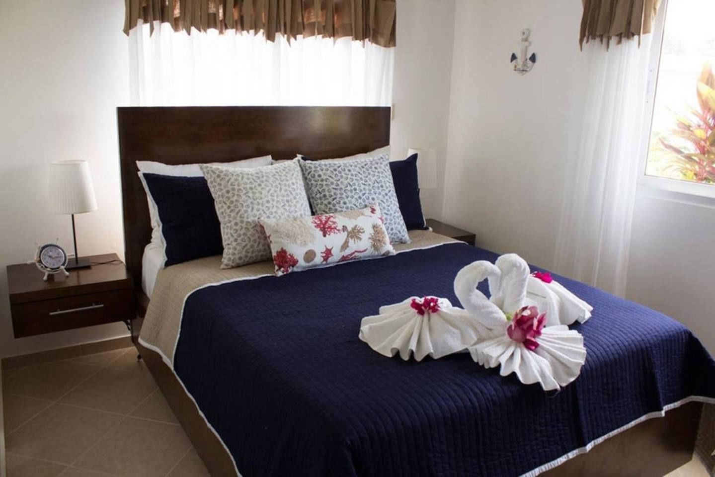 Un lit bien fait, recouvert d’une couvre-lit bleu foncé et agrémenté de coussins décoratifs, une table de chevet avec une lampe et un luminaire fixé au mur.