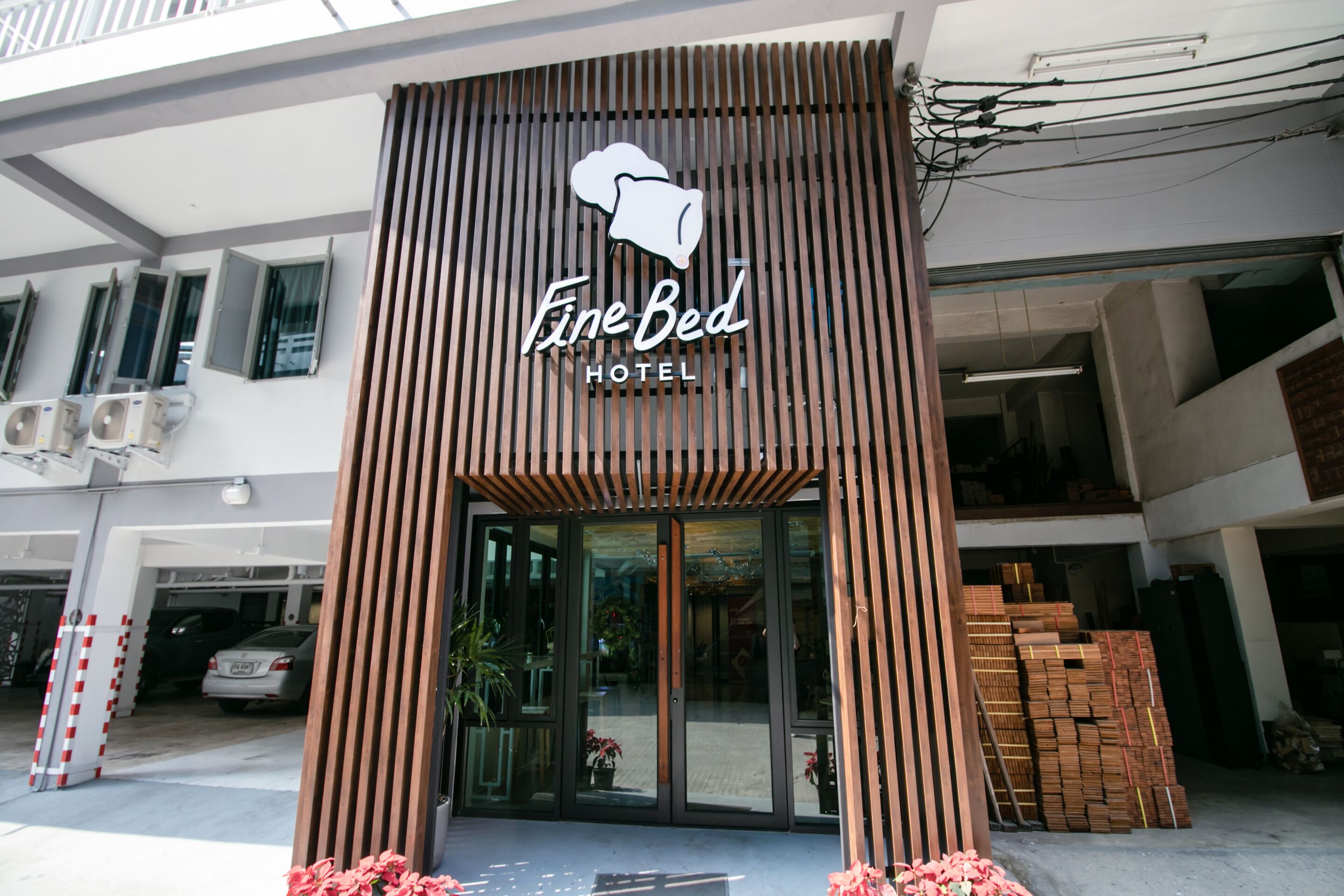 The Fine Bed Hotel entrance with a modern design, including a logo, a covered entrance, and a parking area with cars and construction materials nearby.