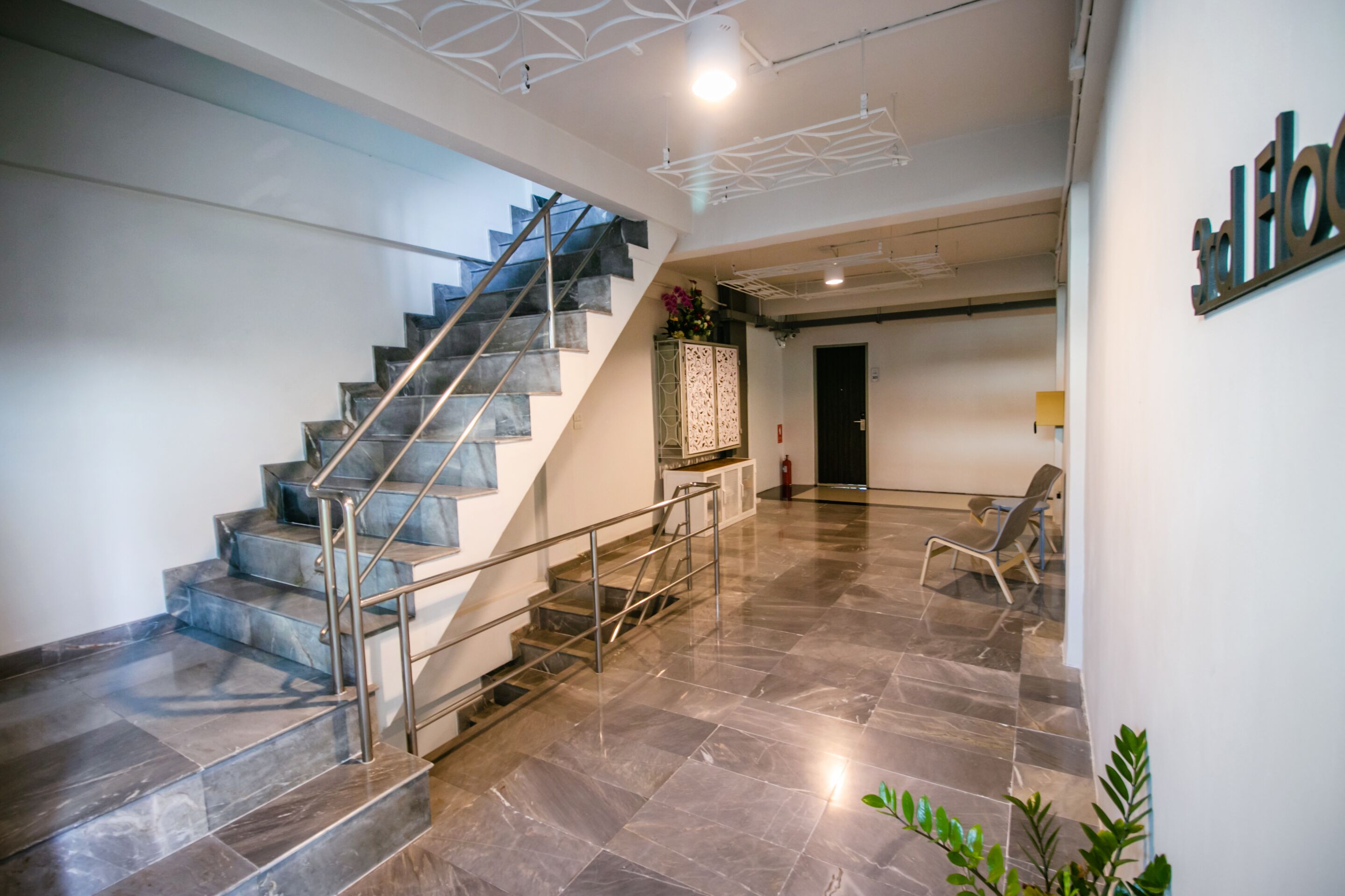 Un interior moderno con una escalera, barandillas metálicas, un mostrador de recepción y una planta.