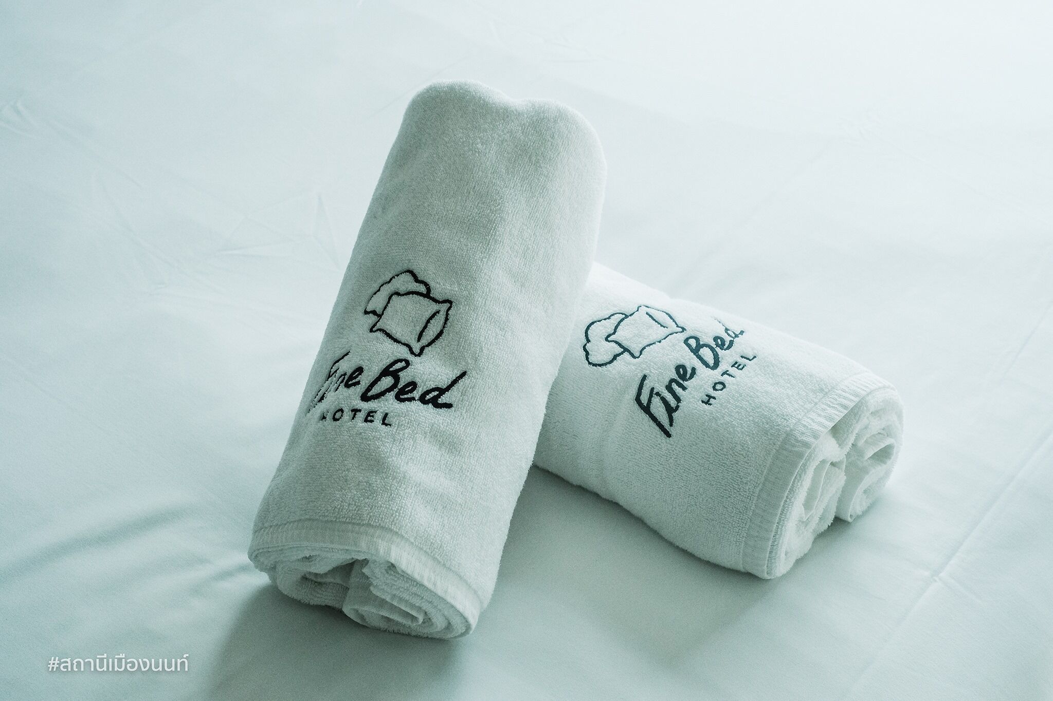Dos toallas blancas enrolladas con texto bordado e icono de cama, sobre una superficie blanca.