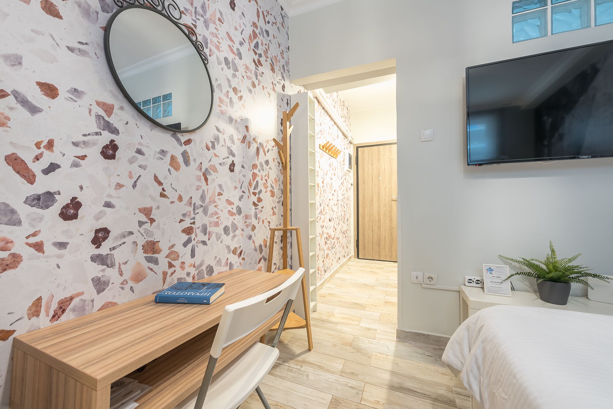 Ein Hotelzimmer mit Terrazzo-Boden, einem Holztisch mit einem Buch, einem Bett, einem Fernseher und einer Pflanze.