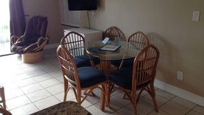 A dining area with a round glass table surrounded by four wicker chairs with blue cushions. A television is mounted on the wall above the table. - Just steps to the Warm Sandy Beach of Southern Florida (Pompano Beach)