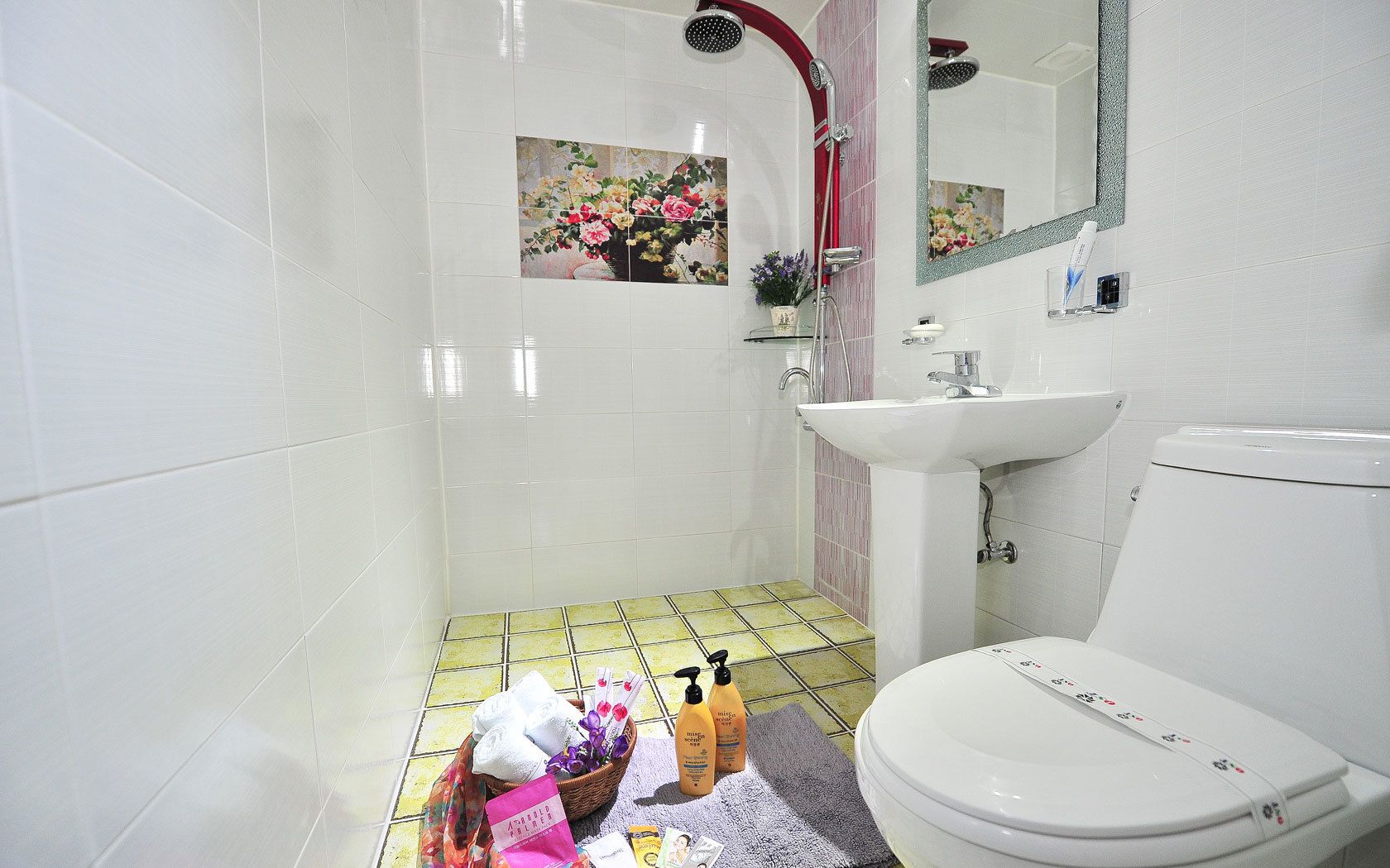 Une salle de bain avec une douche, un lavabo et des toilettes. Un motif floral orne le mur et un panier contenant des articles de toilette est posé sur le sol.