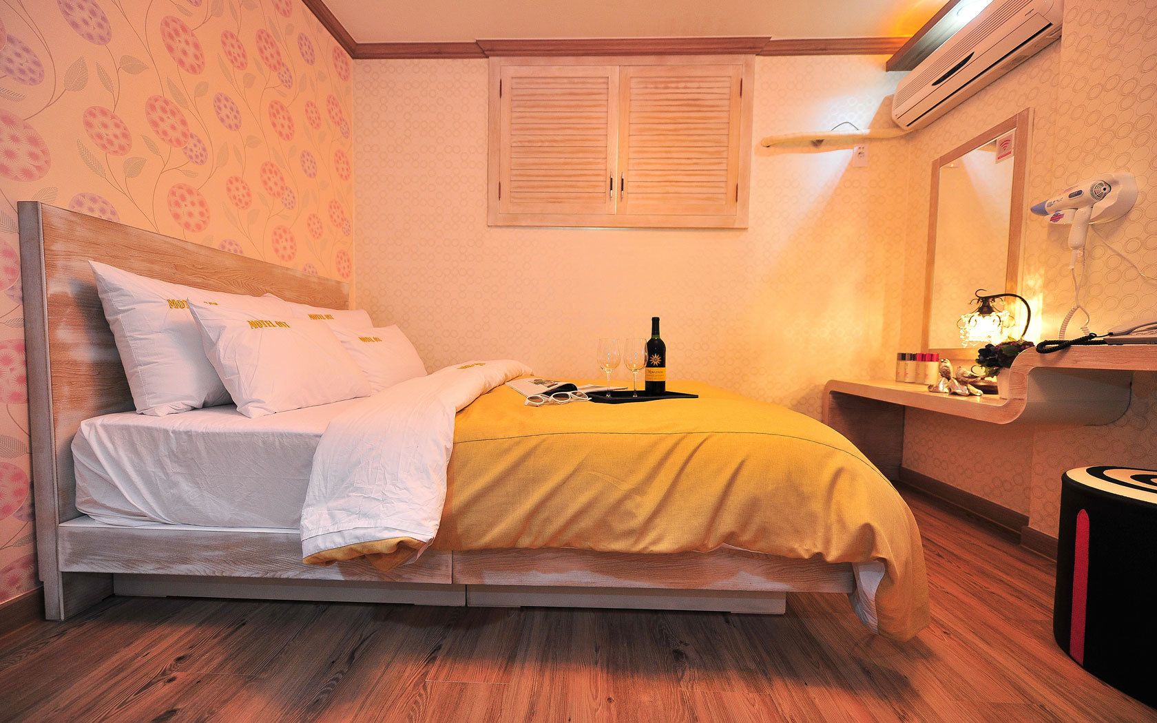 A neatly made bed with a yellow blanket, a bottle of wine, and two glasses on the nightstand.