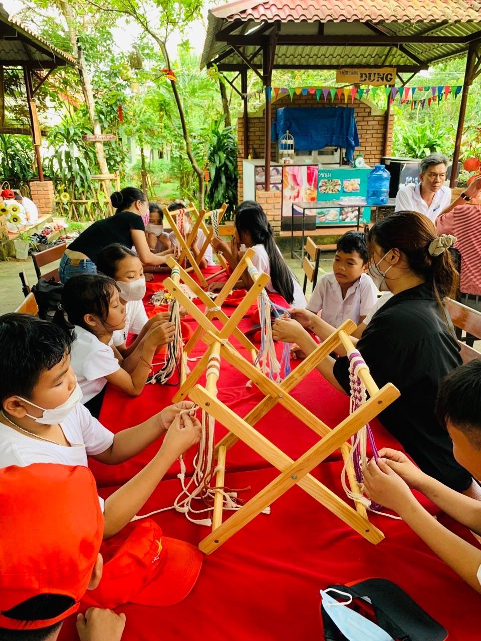 Un groupe de personnes participe à une activité manuelle, assises autour d’une table recouverte d’une nappe rouge, dans un cadre extérieur agrémenté d’une structure en briques et d’une végétation en arrière-plan.