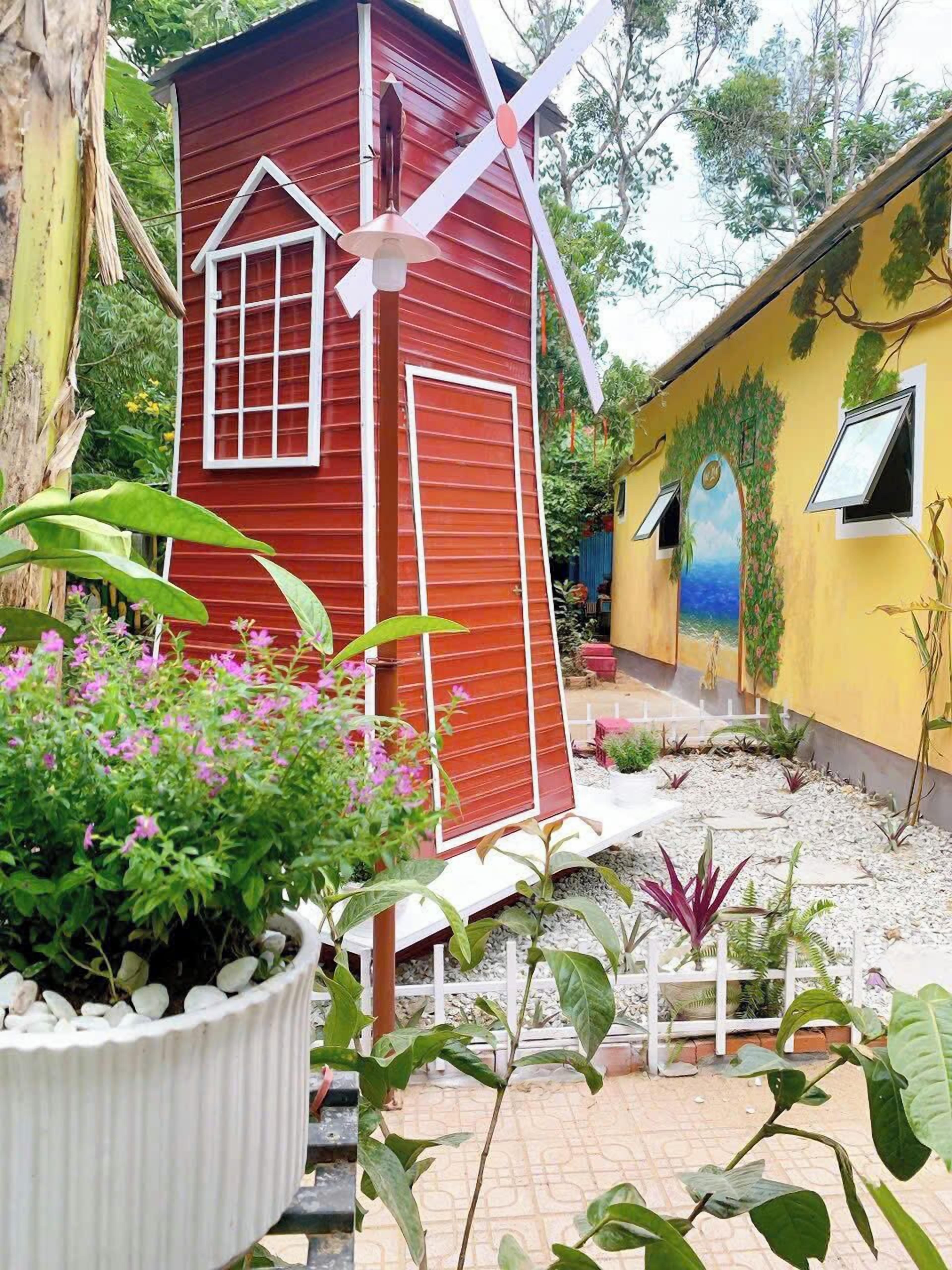 Un edificio rojo y amarillo con una estructura de molino, rodeado de plantas en macetas y un jardín.