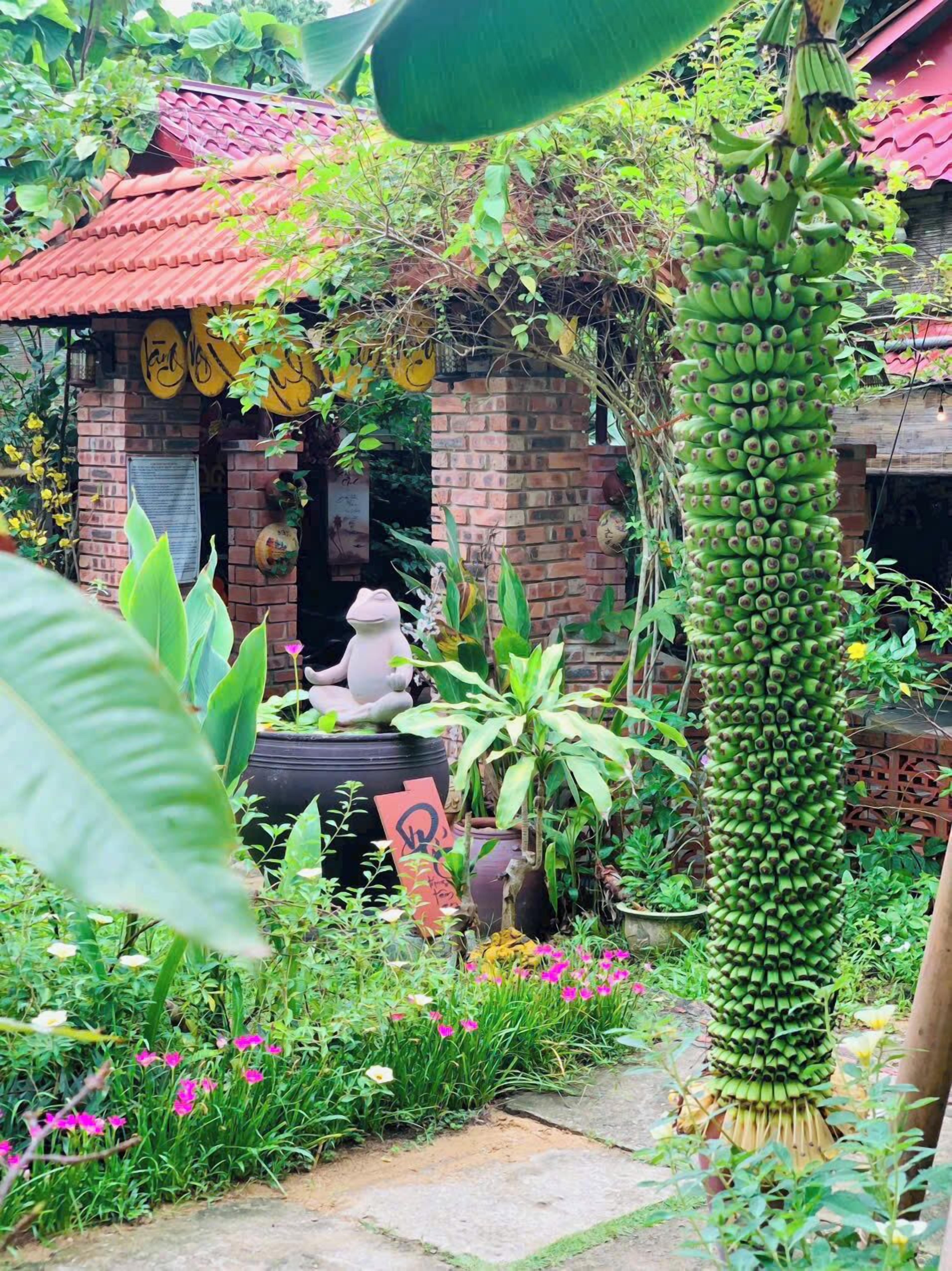 A rustic brick house with a red tiled roof, surrounded by lush greenery and flowering plants.