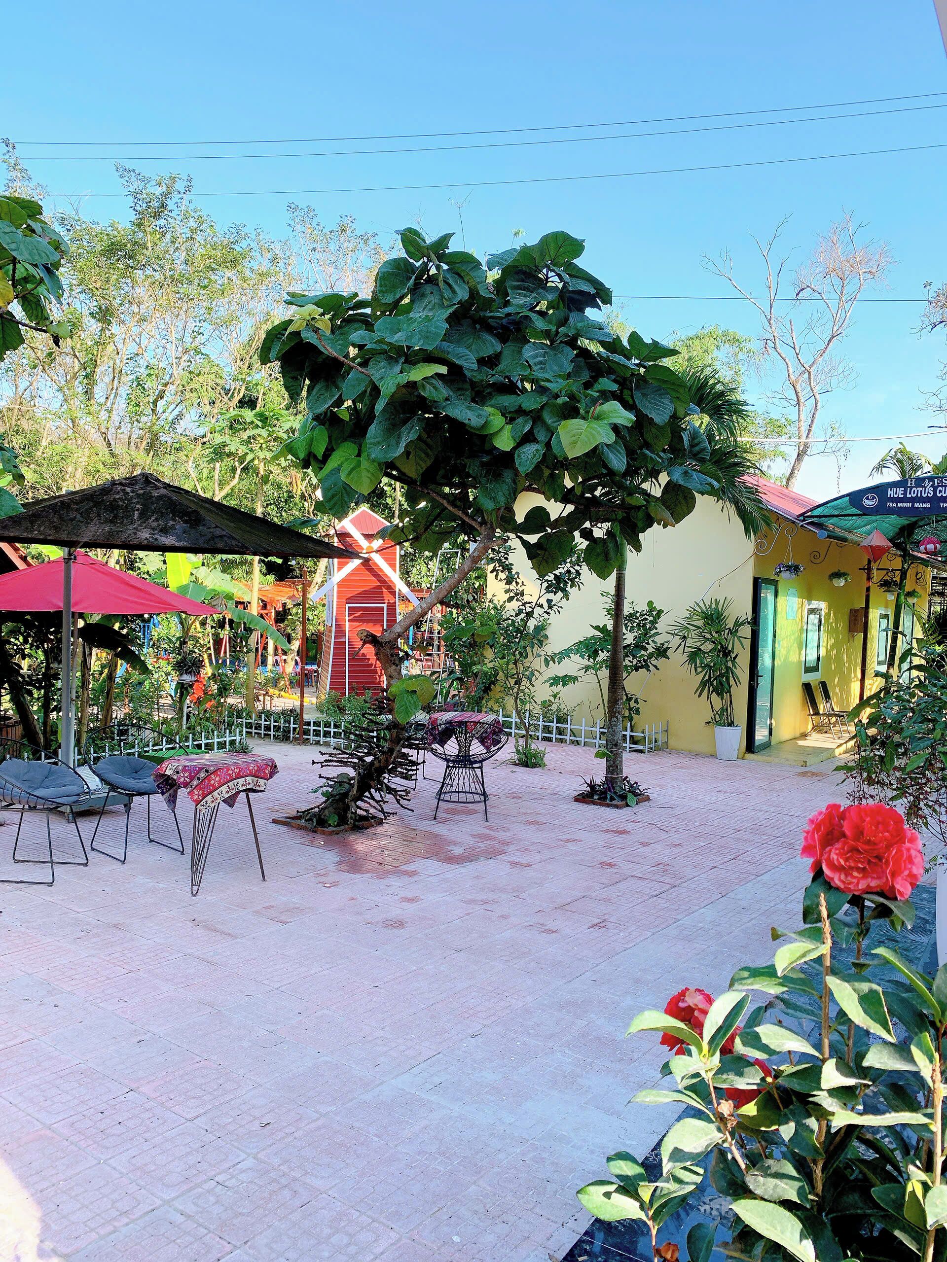 An outdoor seating area with tables and chairs, surrounded by greenery and a building with a yellow facade.