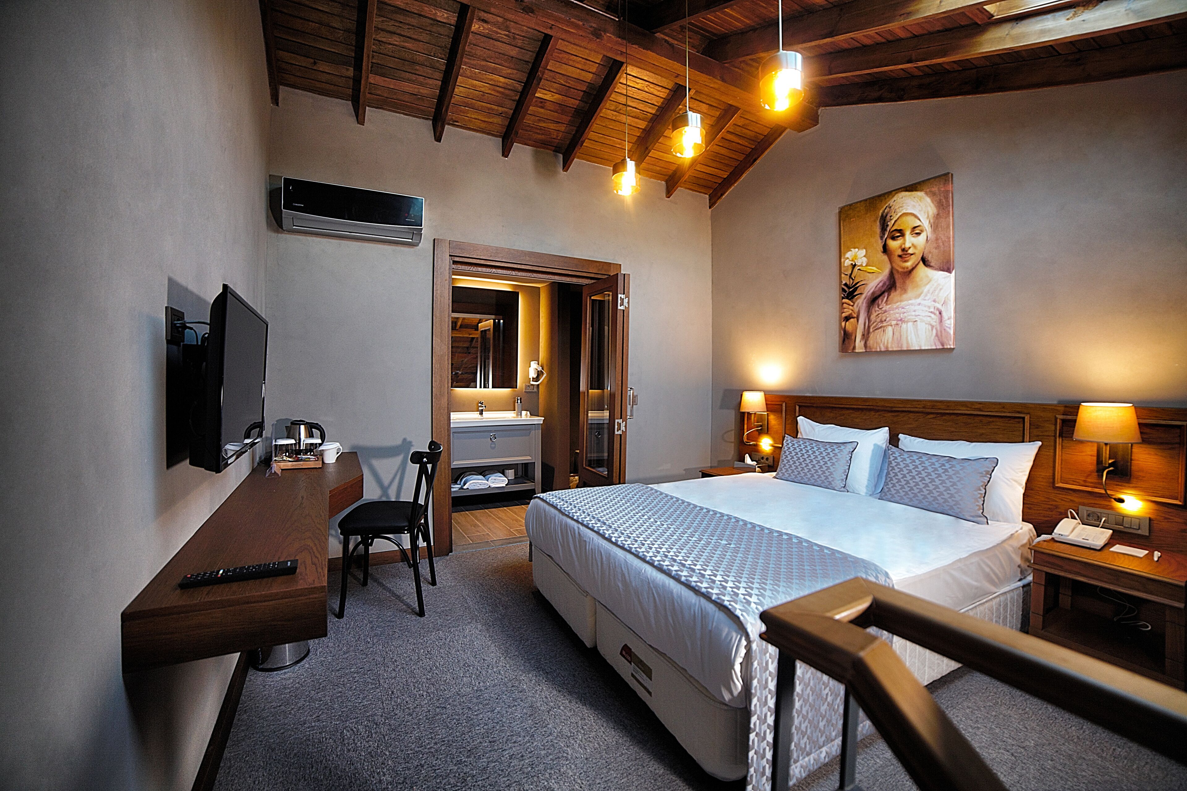 A hotel room with a bed, a desk with a chair, a TV, a bathroom with a mirror, and a painting on the wall.