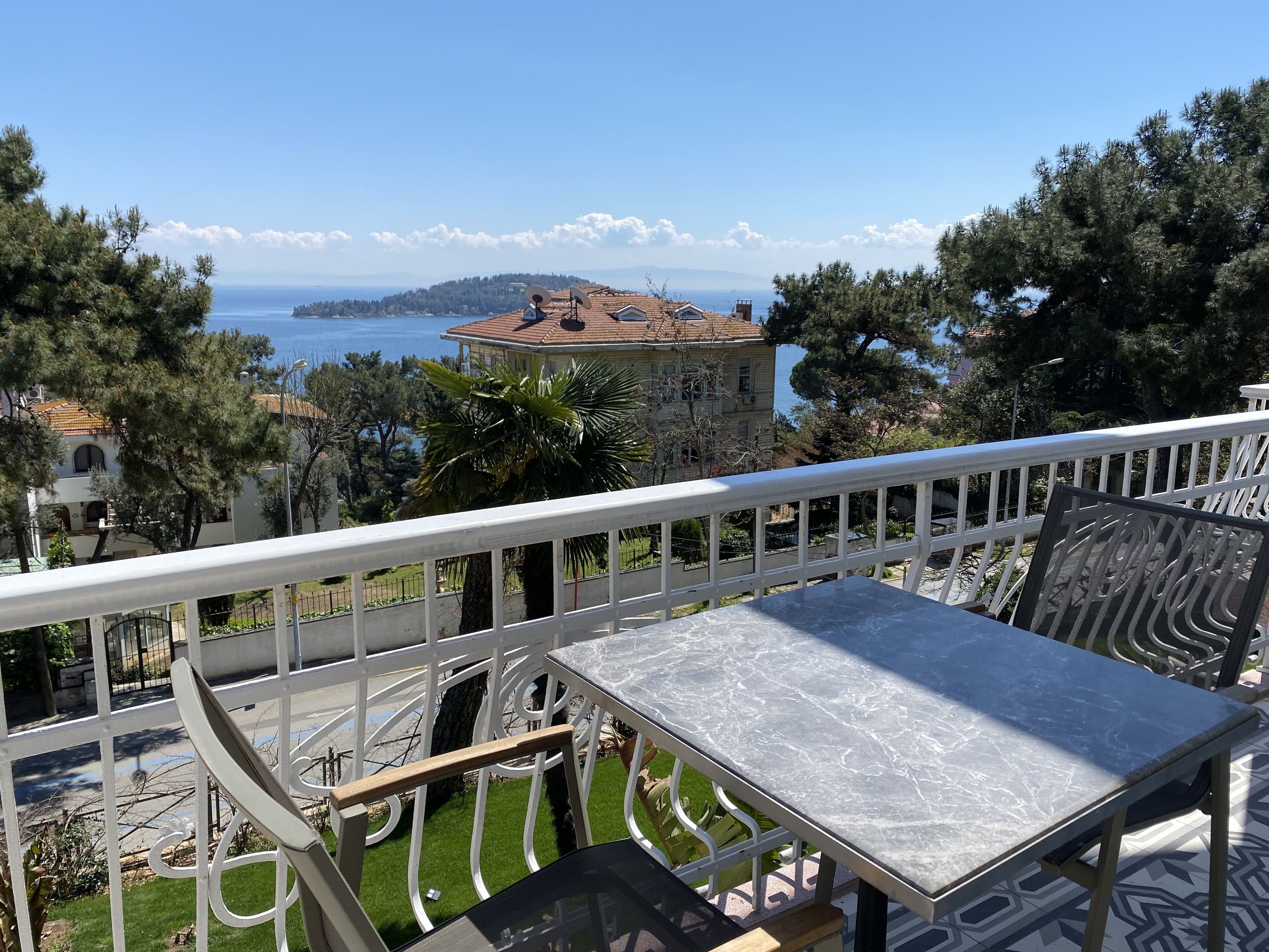 A terrace with a table and chairs, overlooking a coastal view with buildings and trees.