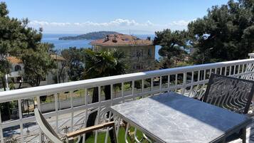 Eine Terrasse mit Tisch und Stühlen, mit Blick auf die Küste, Gebäude und Bäume.