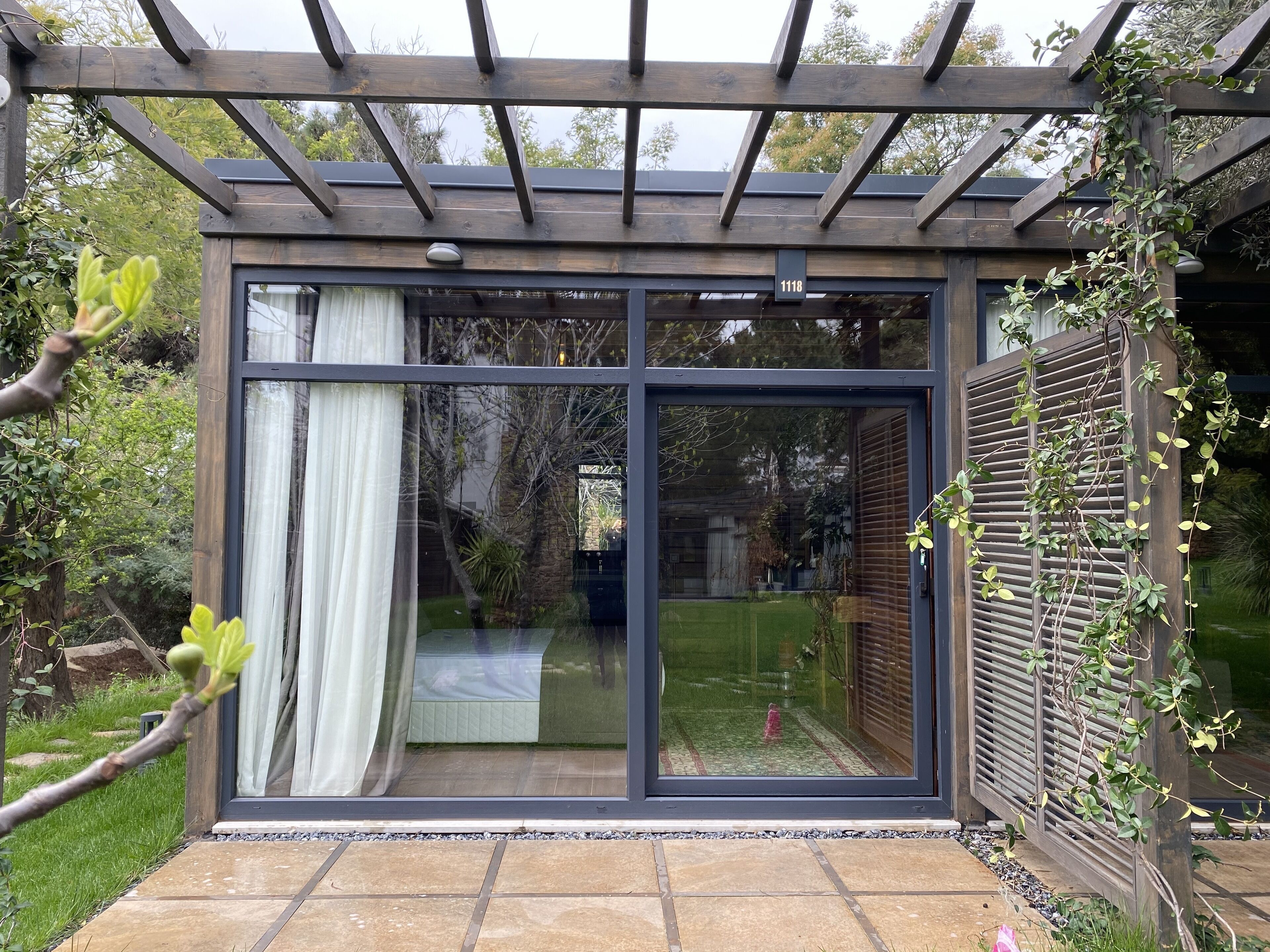 Una entrada moderna con puerta de cristal, una pérgola de madera encima, una placa con número y vista a un jardín.