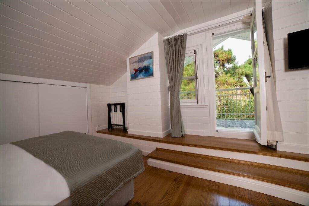 A bedroom with a bed, a window with curtains, a painting on the wall, and a view of greenery outside.