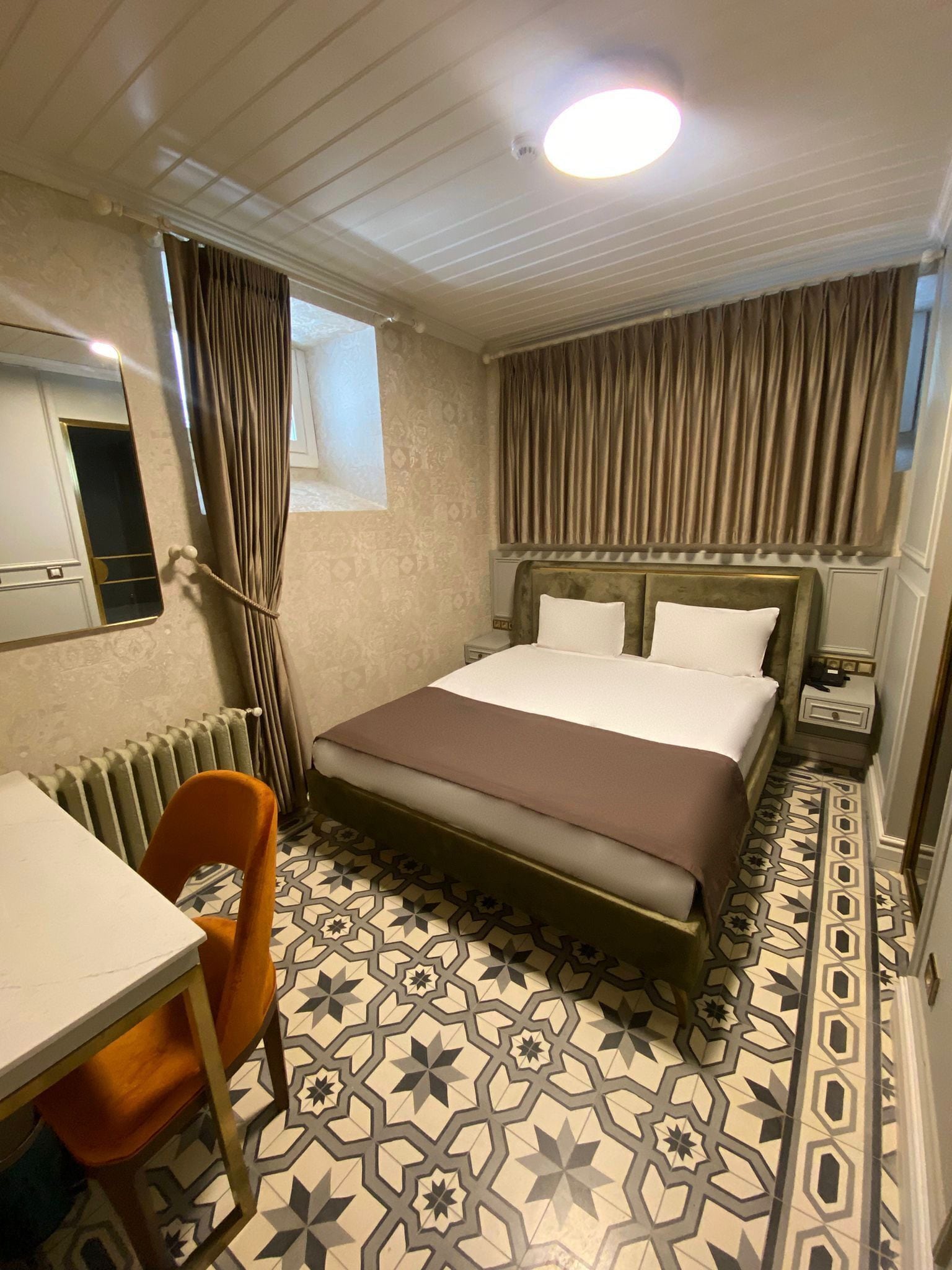 A hotel room with a bed, a desk, a chair, a mirror, and a window with curtains.