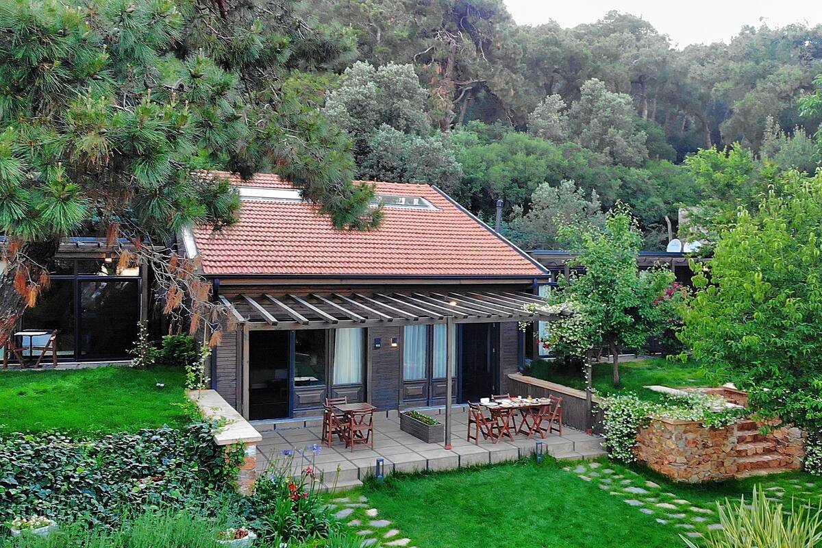 A modern house with a red-tiled roof, a covered patio with outdoor furniture, and a lush garden with various plants and trees.