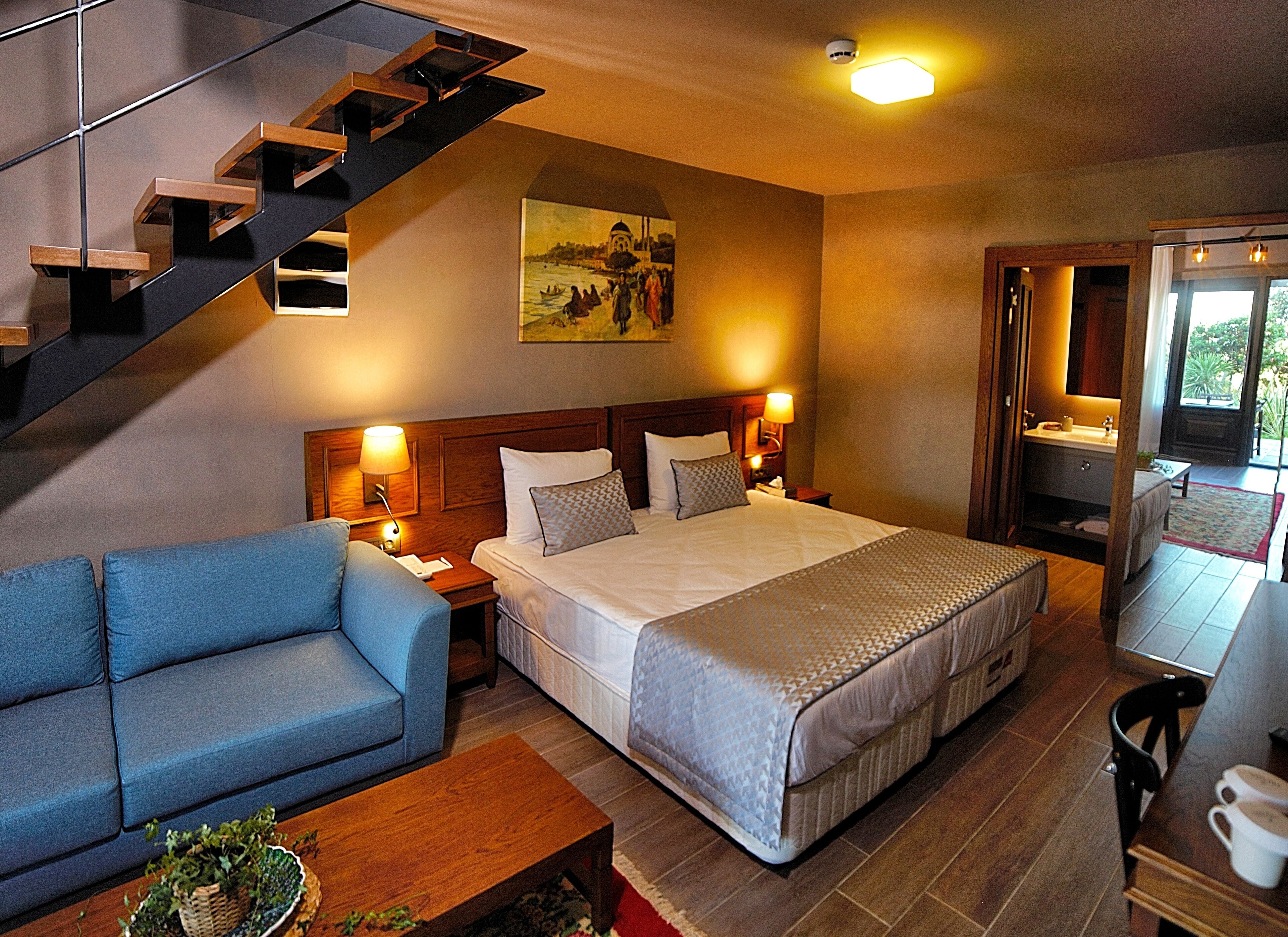 A hotel room with a bed, a sofa, a wooden coffee table, a staircase, a painting on the wall, and a bathroom visible through an open door.