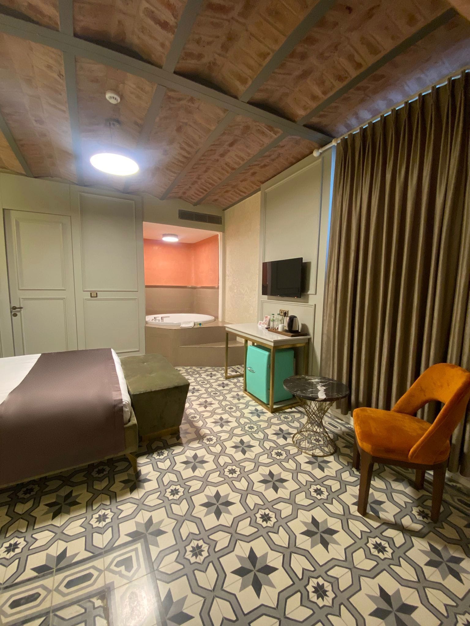 A hotel room with a bed, a small table, a chair, a television, and a bathroom visible through an open door.