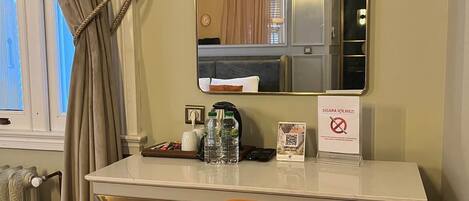 A hotel room with a white desk, a mirror, a chair, a radiator, and a window with curtains.