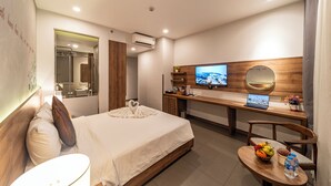 Habitación de hotel con cama, escritorio, silla, televisor y una mesita con frutero y botella de agua.