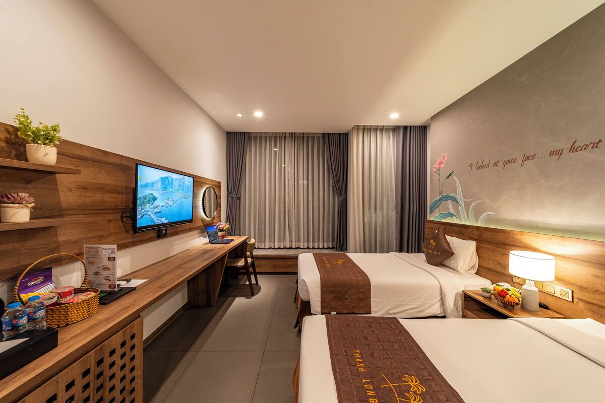 A hotel room with two beds, a desk with a computer, a TV, and a wall with a scenic painting.