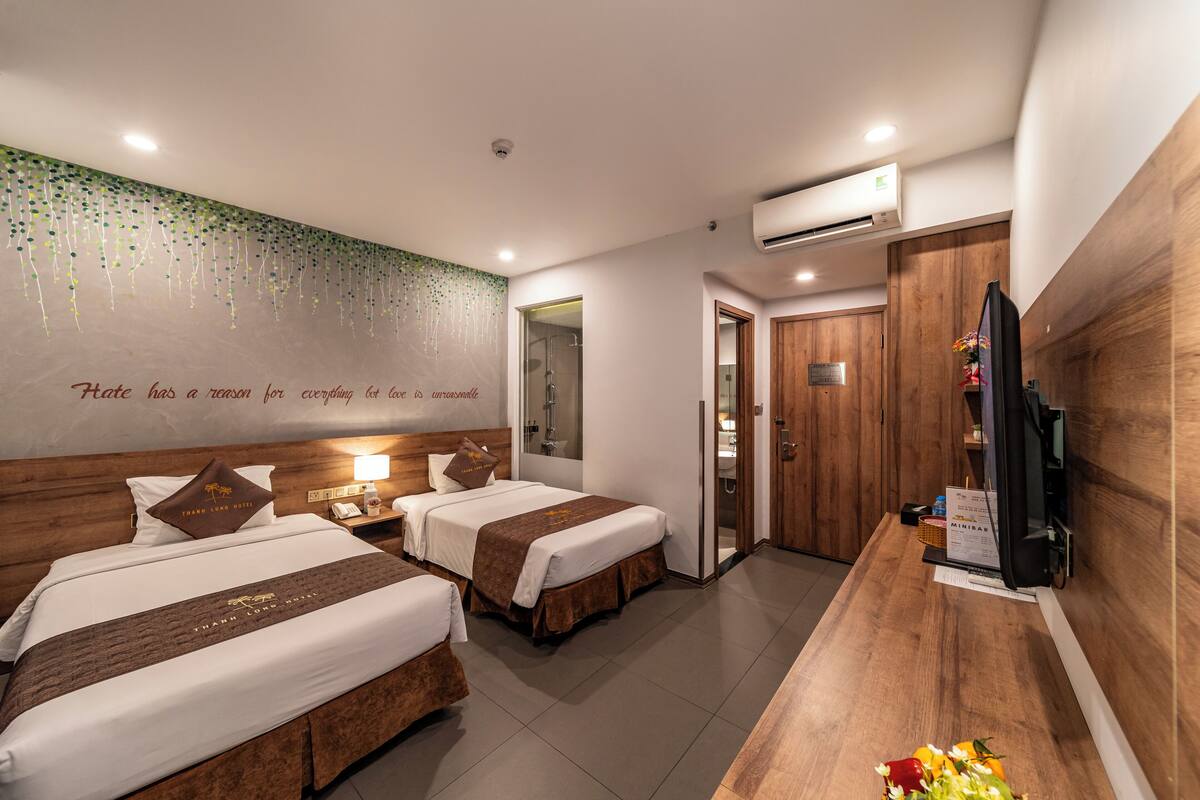 A hotel room with two beds, a wooden desk, a television, and a wall with a quote.