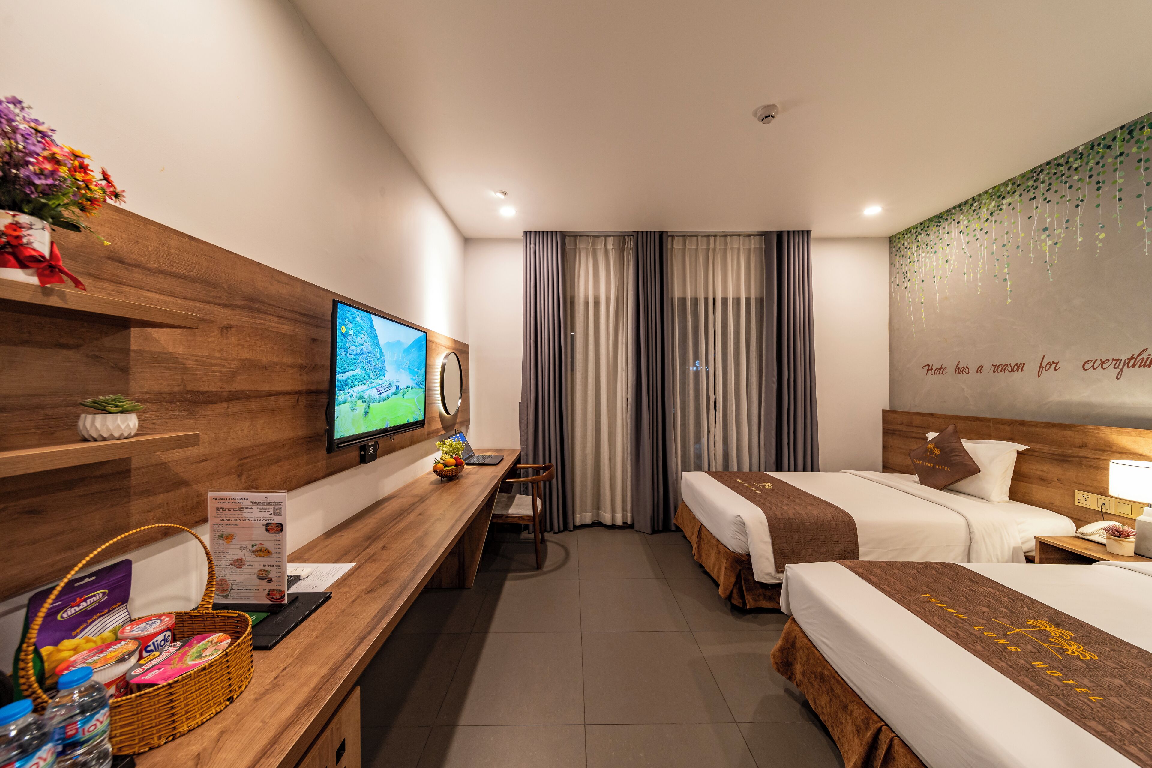 A hotel room with two beds, a wooden desk with a TV, a small table, and a wall with a quote.