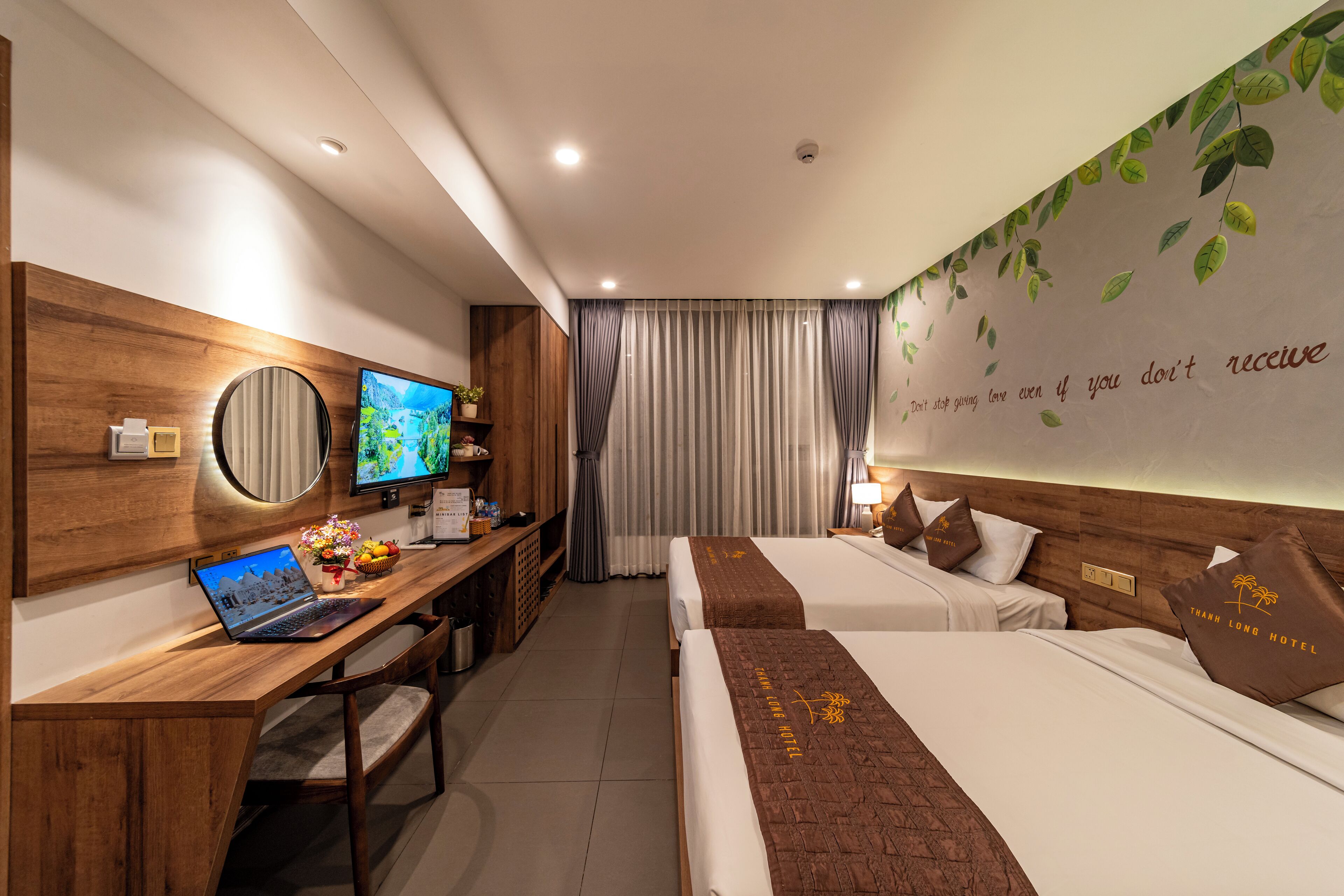 Een hotelkamer met twee bedden, een bureau met een laptop, een televisie en een muur met een inspirerende quote.