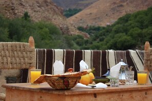 Desayuno con una cesta tejida, jugo de naranja y una tetera sobre una mesa de madera, con montañas al fondo.