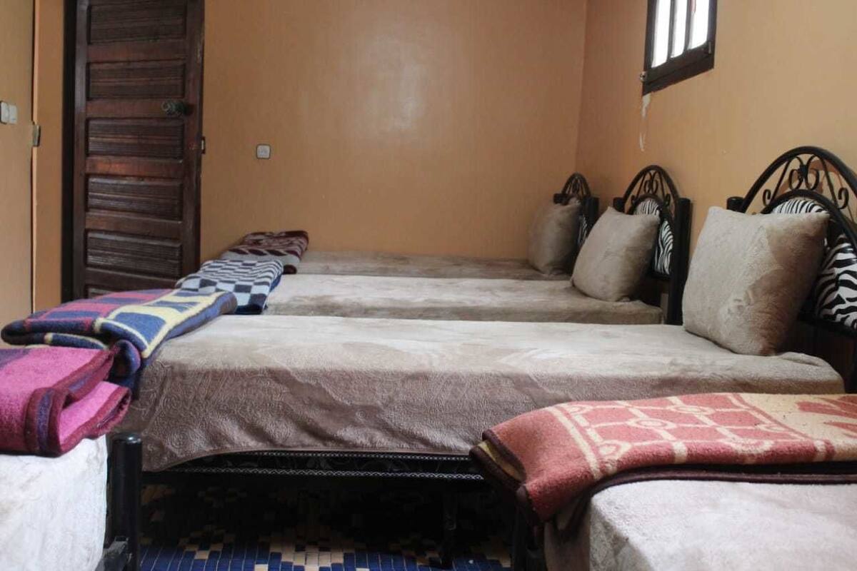 Ein Zimmer mit zwei Betten, jeweils mit einem gemusterten Überwurf und Kissen. Eine Holztür und ein kleines Fenster sind ebenfalls vorhanden.