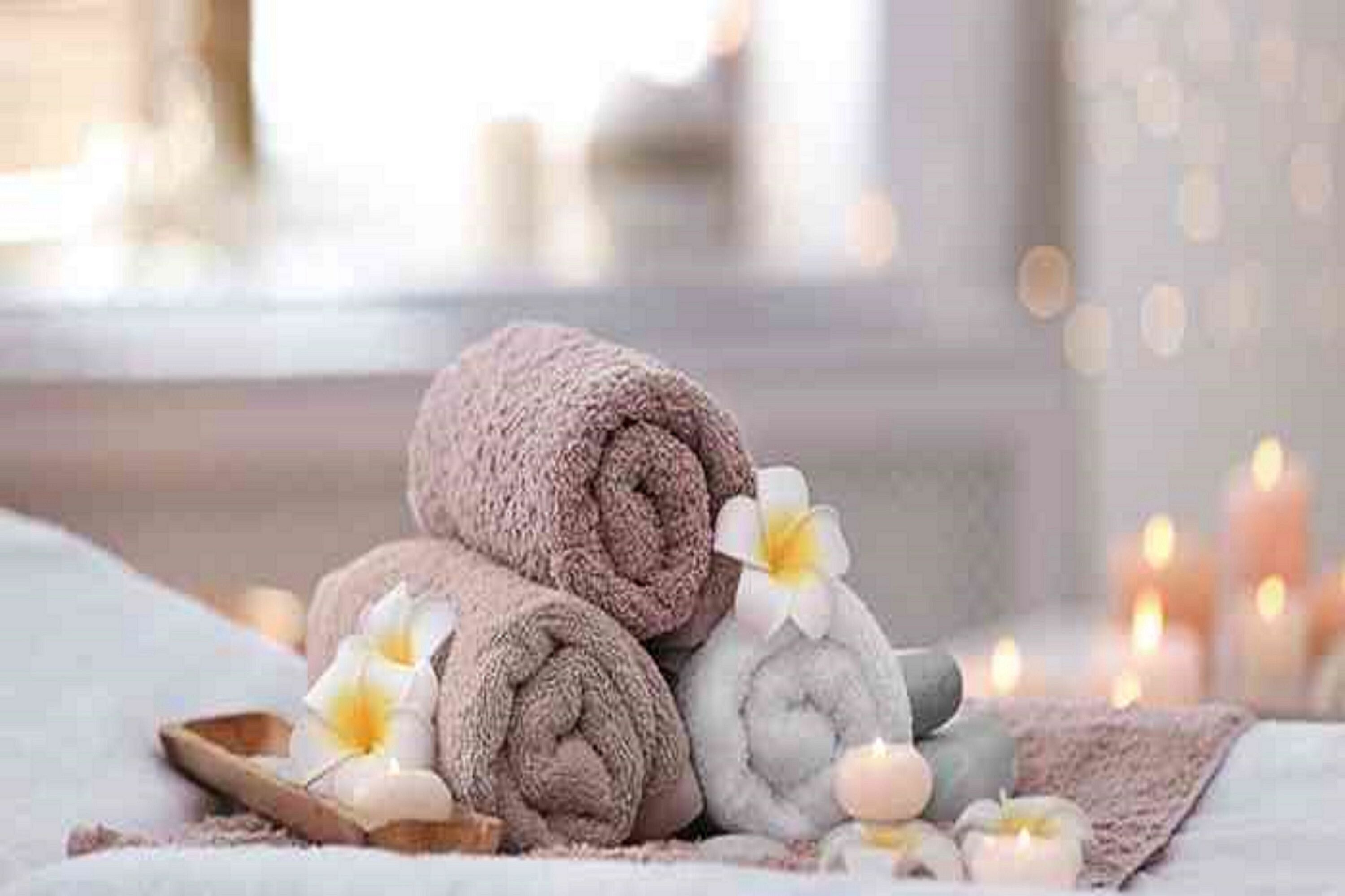 A stack of neatly rolled towels, a small tray, and candles on a bed with a soft, neutral-colored blanket.