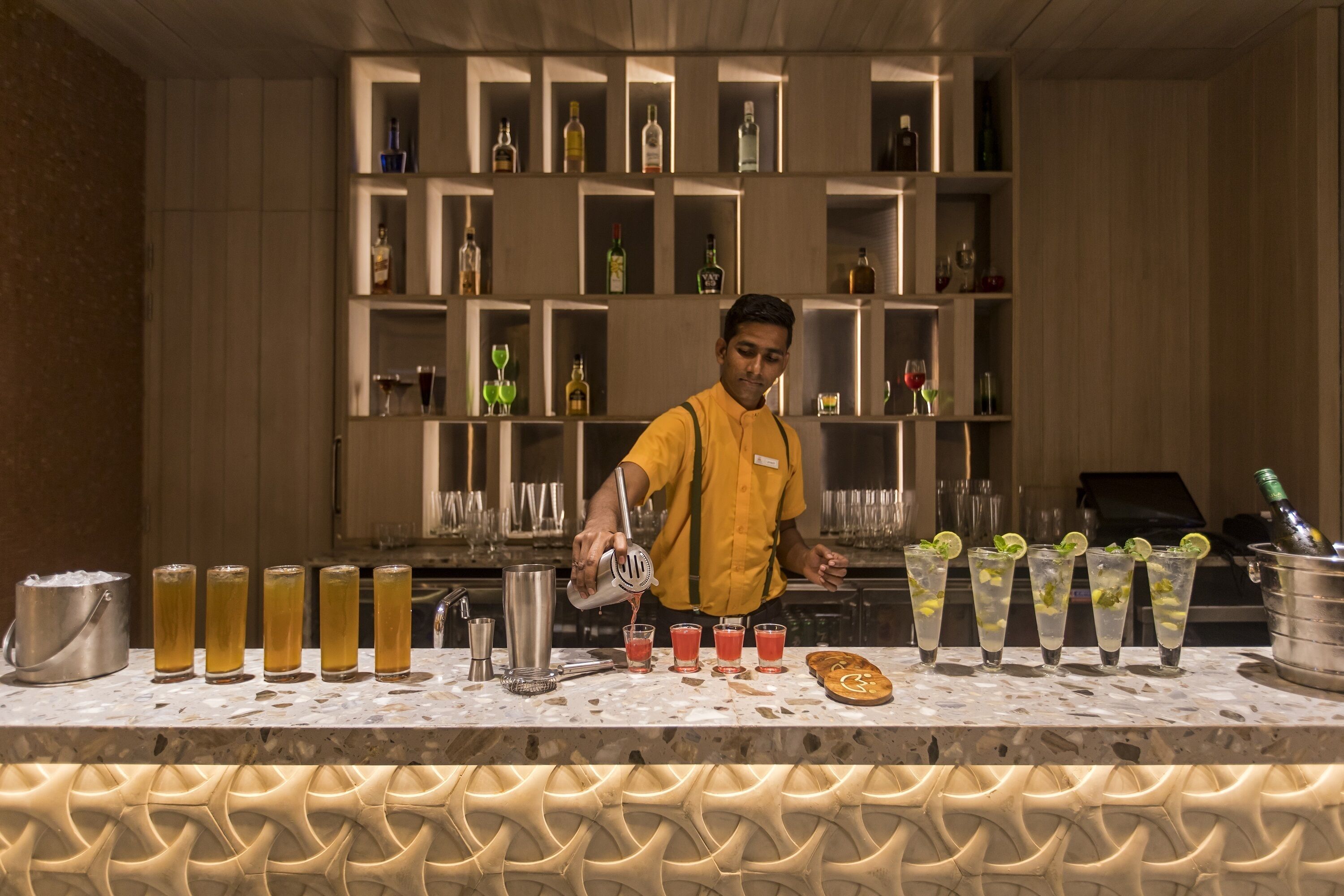 Zona de bar con un bartender sirviendo bebidas. En el fondo, estanterías con botellas y vasos.