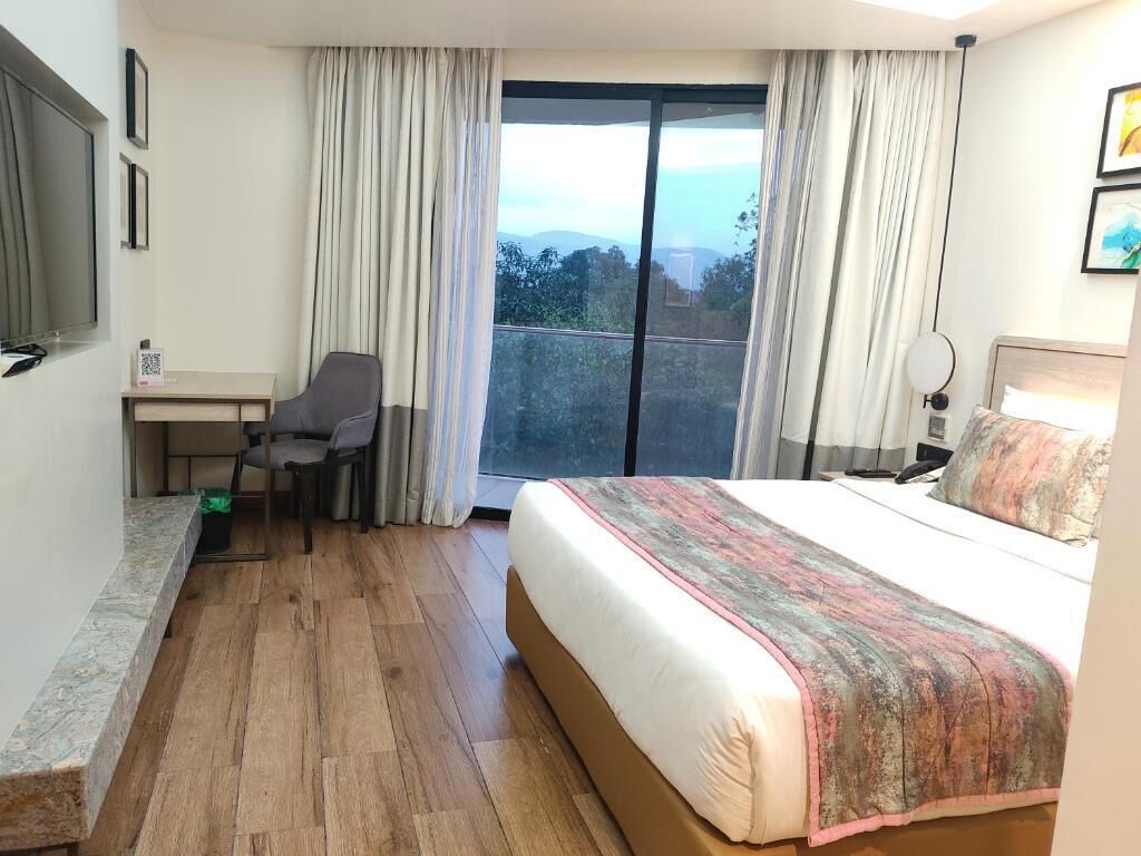 A hotel room with a large bed, a desk with a chair, a window with curtains, and a view of the outdoors.