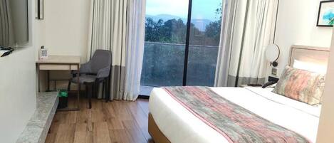 A hotel room with a large bed, a desk with a chair, a window with curtains, and a view of the outdoors.