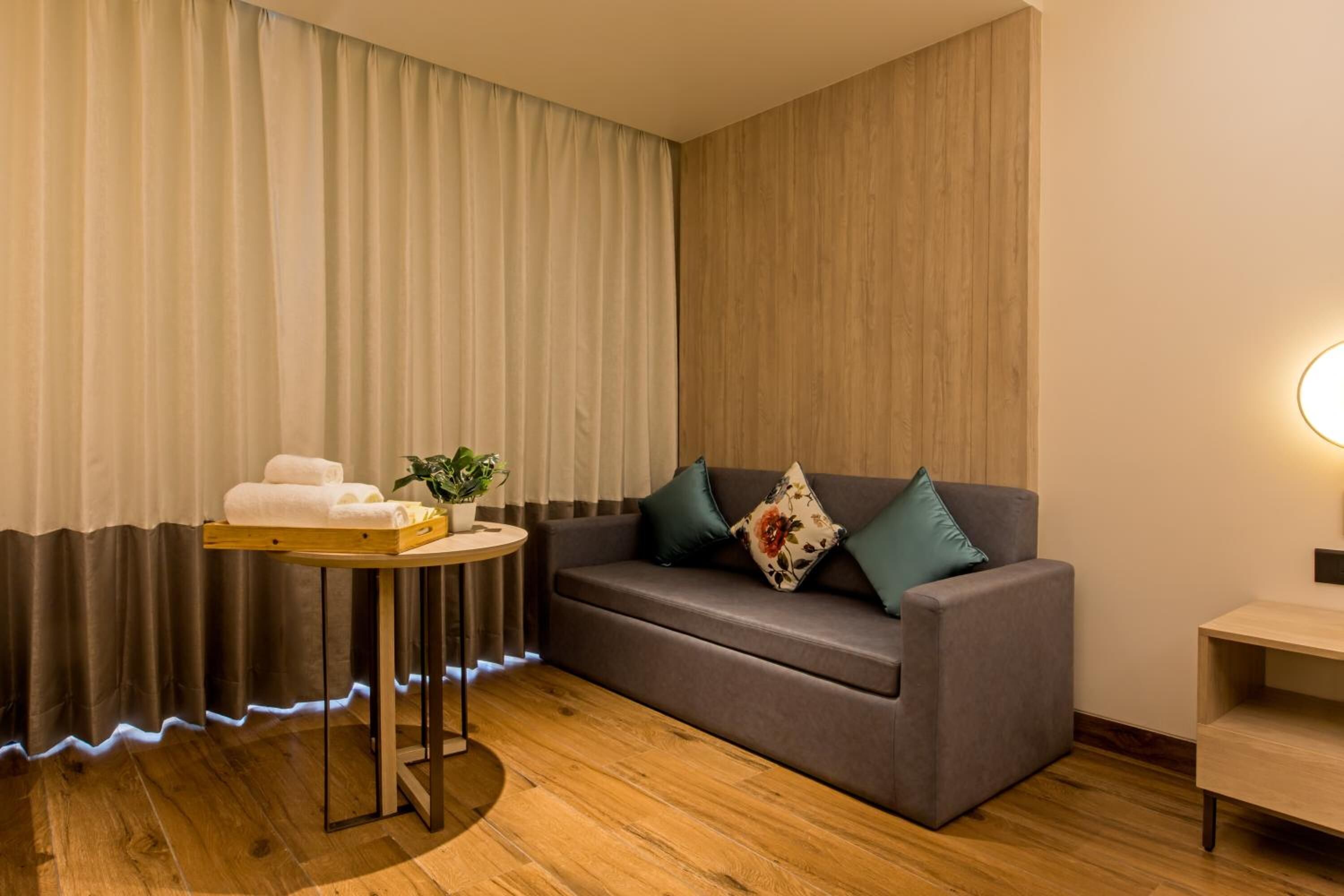A modern room with a grey sofa, a small round table with towels and a plant, and a wooden floor.