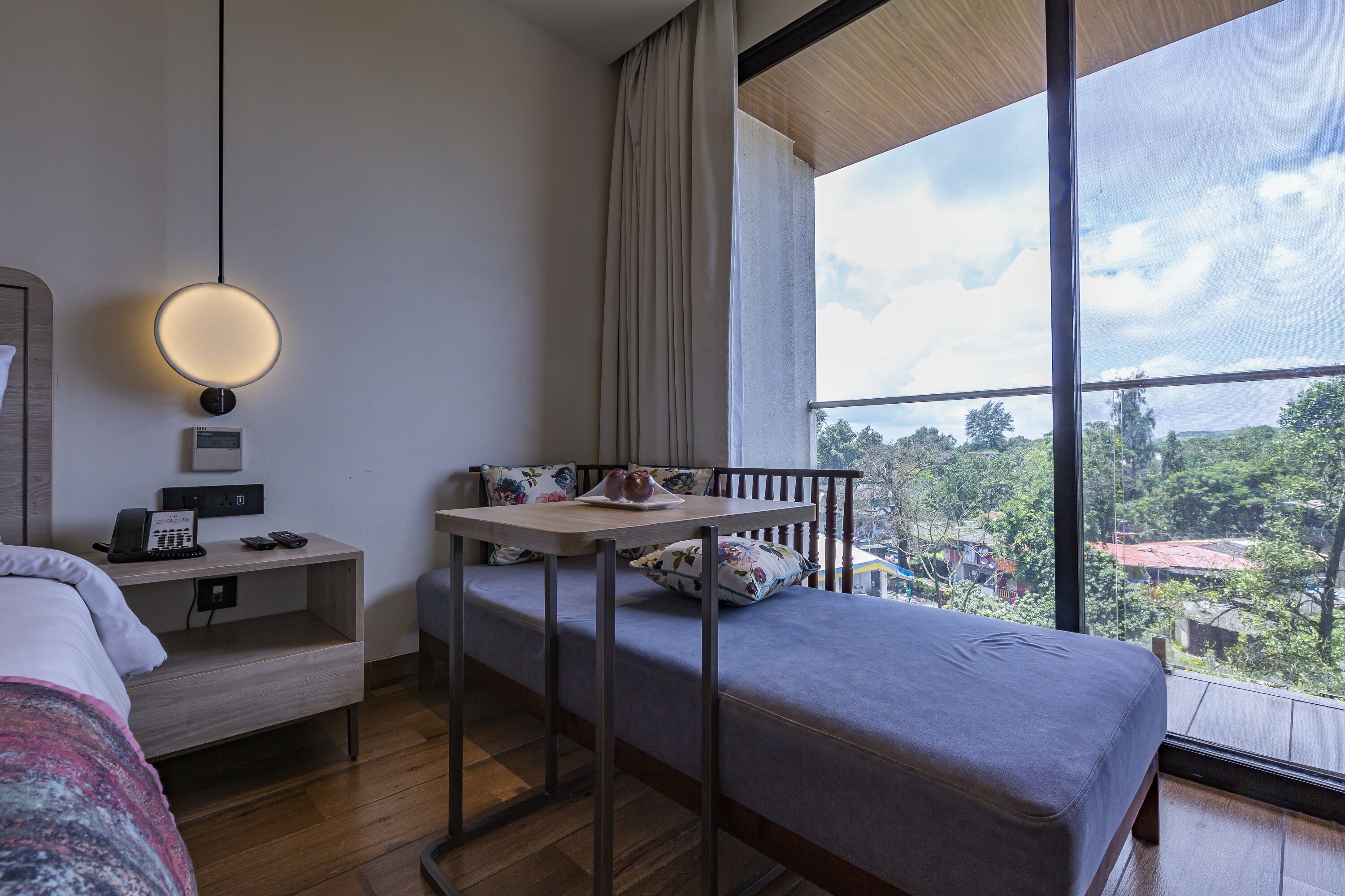 A hotel room with a bed, a desk with a lamp, a phone, and a view of the city.