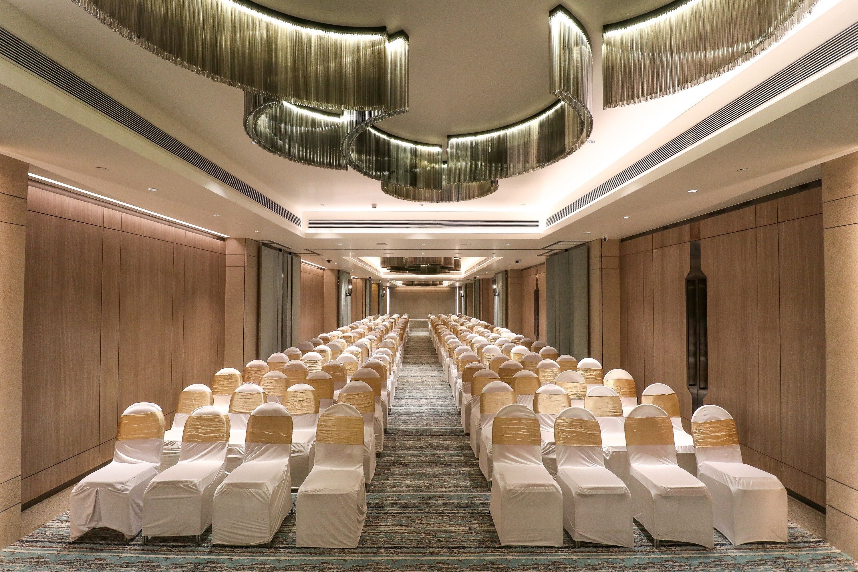 A hotel conference room with rows of chairs covered in white tablecloths and gold sashes, a patterned carpet, and a modern ceiling design.