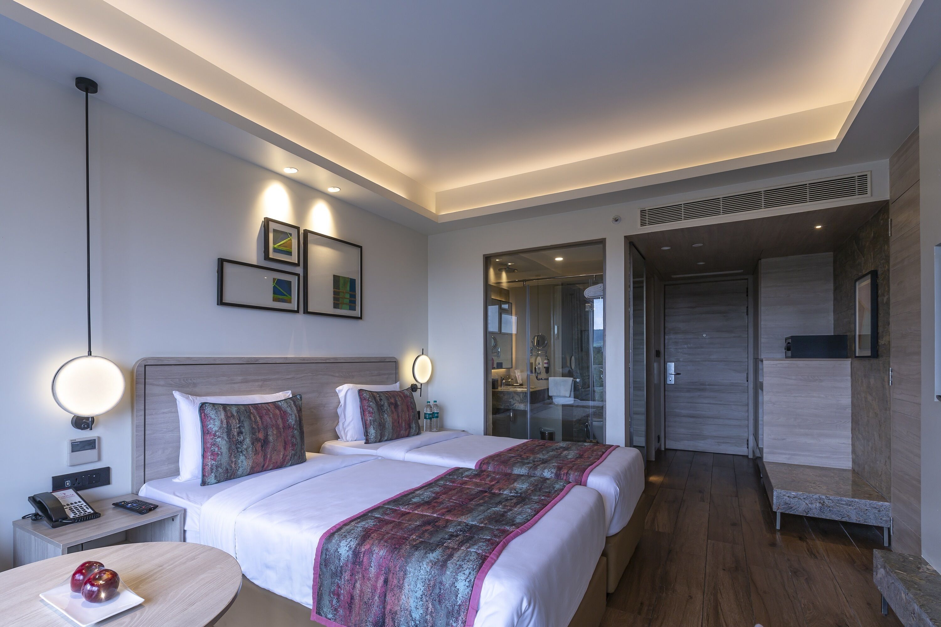 A modern hotel room with two beds, a bedside table with a phone, a round table with fruit, and a bathroom visible through an open door.
