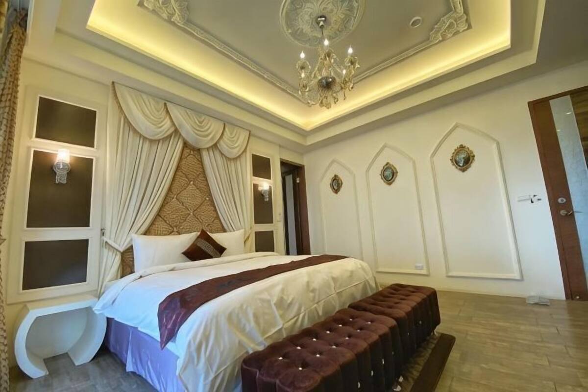A bedroom with a large bed, a bench, a chandelier, and decorative wall panels.