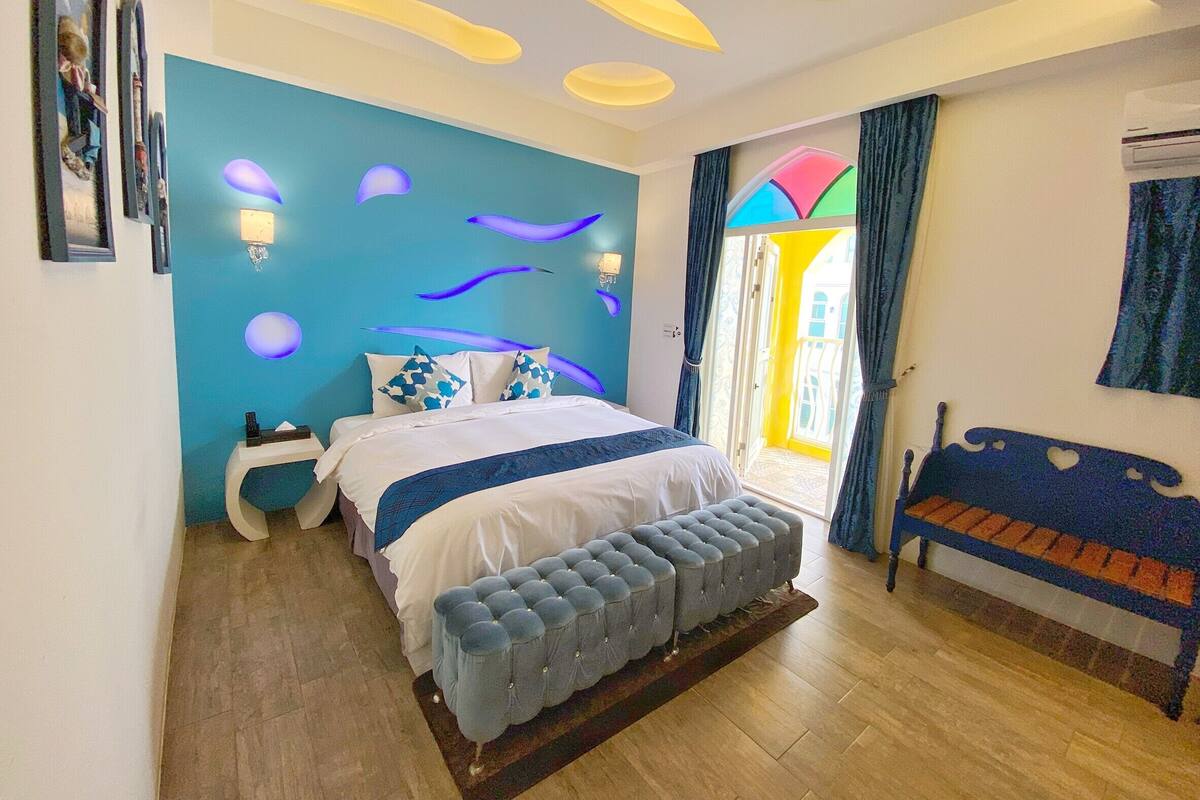 A bedroom with a bed, a bench, a wall with light fixtures, and a door leading to a balcony.
