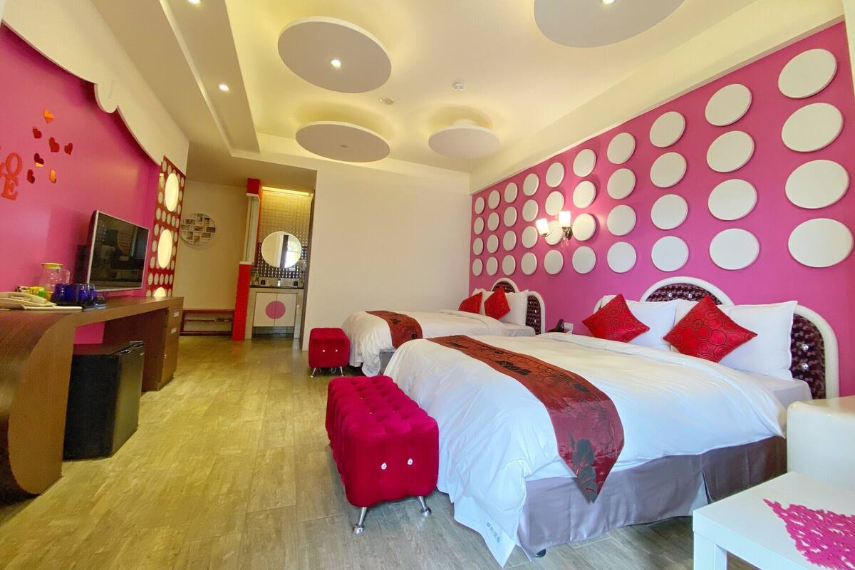 A hotel room with a pink and white color scheme, a large bed with red and white bedding, a desk with a computer, and a vanity with a mirror.