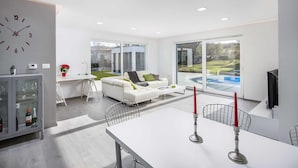 A modern living room with a white sofa, a dining table with two candles, and large windows overlooking a pool. - Villa Aida (Labin)