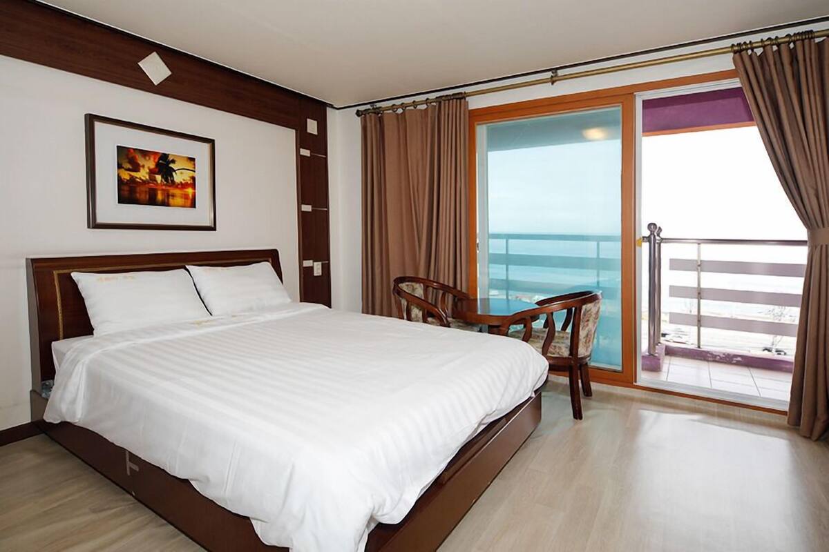 Habitación de hotel con una cama grande, una silla, un balcón con vista al mar y un cuadro enmarcado en la pared.