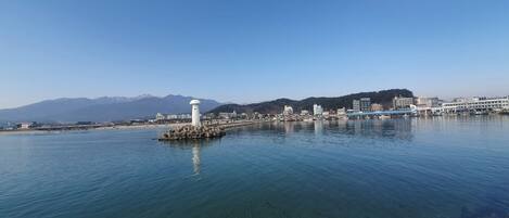 A coastal cityscape with a prominent white lighthouse on a small island, clear blue water, and a backdrop of mountains.