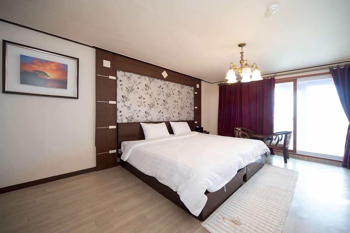 Un dormitorio moderno con una cama grande, cabecera de madera, un cuadro enmarcado, una araña y vistas al exterior.